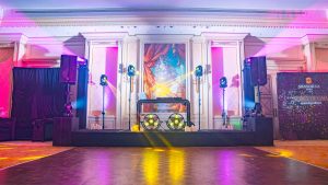grande salle habillée par un plafond de guirlandes LED 2700K en nappe, uplights discrets aux murs, zone DJ cadrée par deux totems autoportés.